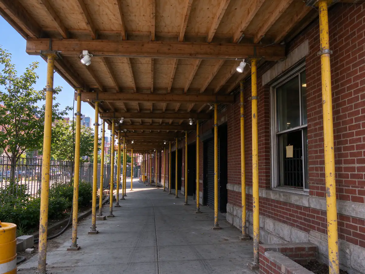 A sidewalk bridge with a plywood ceiling and lights over a city sidewalk in front of a brick building
